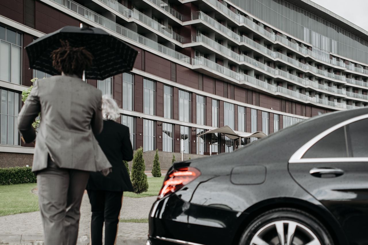 Two professionals in suits under an umbrella walking towards a modern building beside a luxury car.
